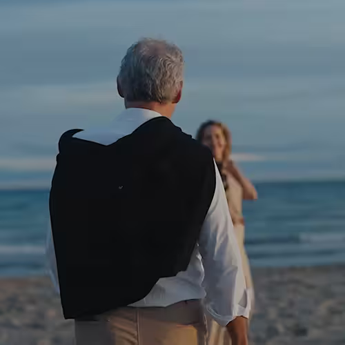 Older man walking on beach toward smiling woman by the ocean at sunset.