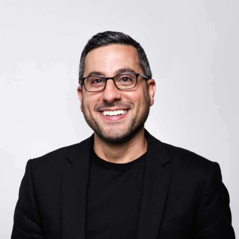 Smiling man with glasses and dark hair wearing a black blazer and shirt against a gray background.