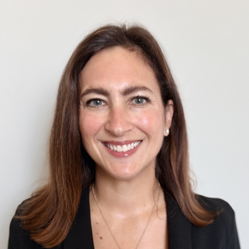 Smiling woman with medium-length brown hair wearing a black top and pearl earrings against a white background.