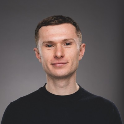 Headshot of a young man with short brown hair wearing a black crewneck shirt against a gray background.