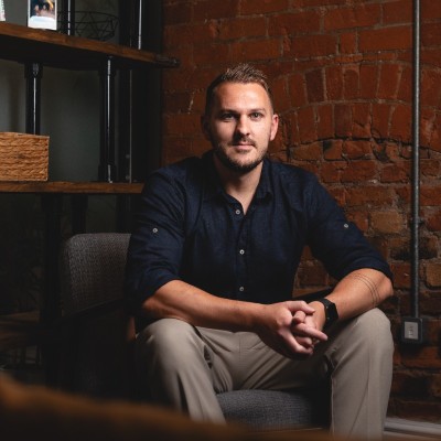 Man with short hair and beard sitting on a chair against a brick wall, hands clasped.