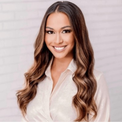 Smiling woman with long wavy hair wearing a white blouse in front of a light brick wall.