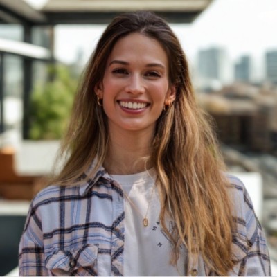 Smiling young woman with long hair wearing a plaid shirt and gold necklace in an outdoor urban setting.