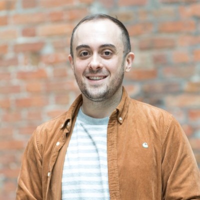 Smiling man with short hair in a tan jacket and striped shirt standing against a brick wall.