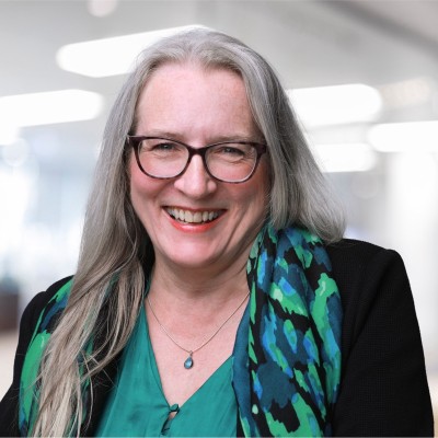 Smiling woman with glasses, long gray hair, green blouse, and blue-green patterned scarf on blurred background.