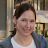 Smiling woman with dark hair wearing earrings and a blue top in a warmly lit indoor setting.