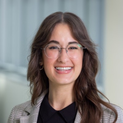 Smiling woman with long brown hair and glasses wearing a plaid blazer and black top.