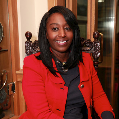 Smiling woman with straight black hair wearing a red blazer and sitting in a carved wooden chair.