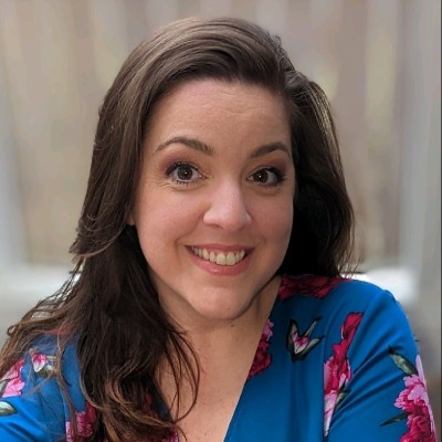 Smiling woman with long brown hair wearing a blue top with pink floral patterns.