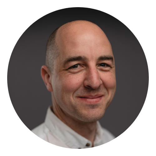 Smiling bald man with light skin and brown eyes wearing a white shirt on gray background.