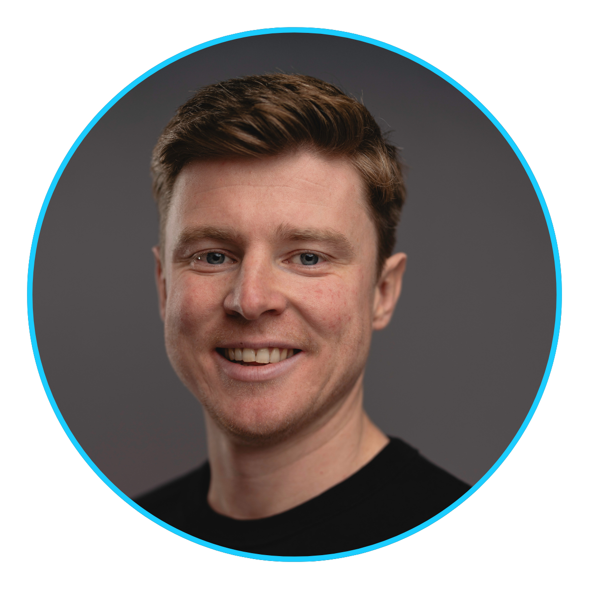 Smiling young man with short brown hair and blue eyes wearing a black shirt on gray background.