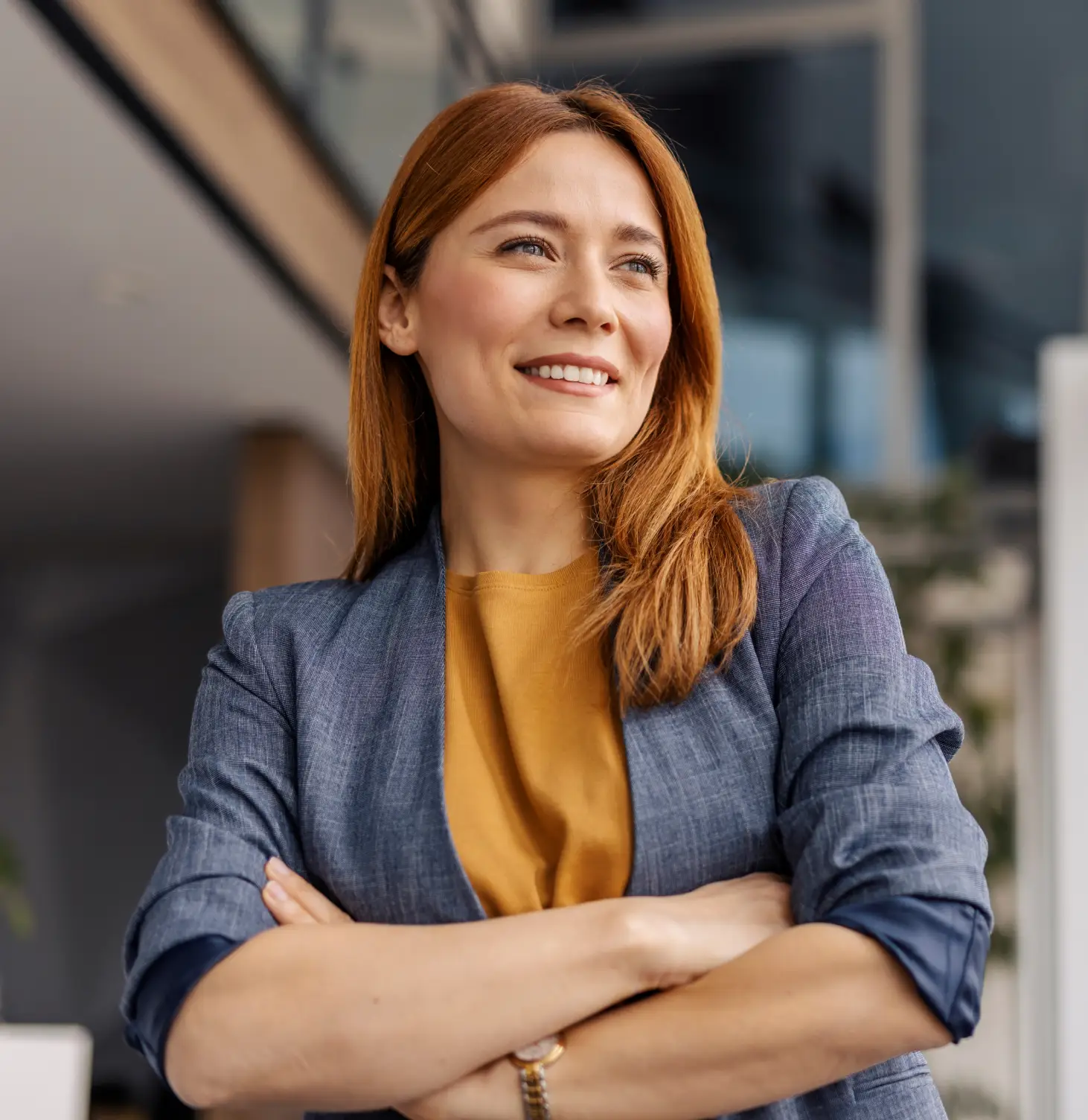 Confident woman with red hair smiling and crossing arms, wearing a gray blazer and mustard shirt.