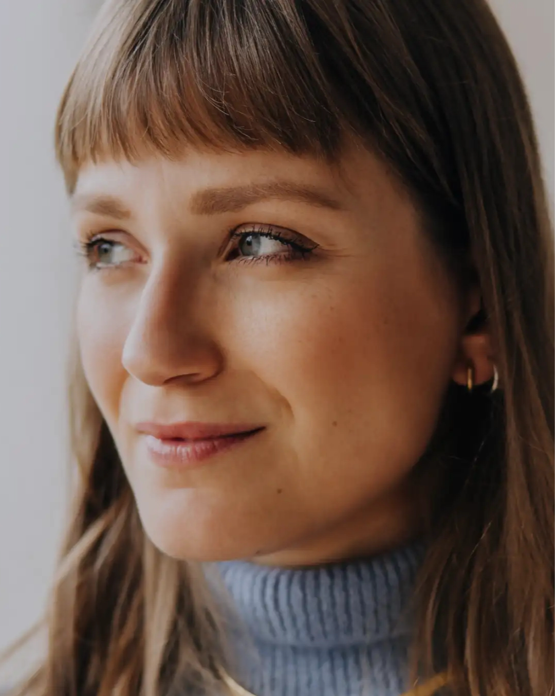 Close-up of a woman with brown hair and blue eyes wearing a blue turtleneck, looking left and smiling slightly.