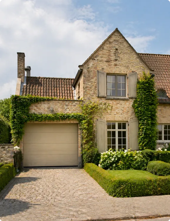 Maison en pierre avec garage, fenêtres à volets et plantes grimpantes sous un ciel partiellement nuageux.
