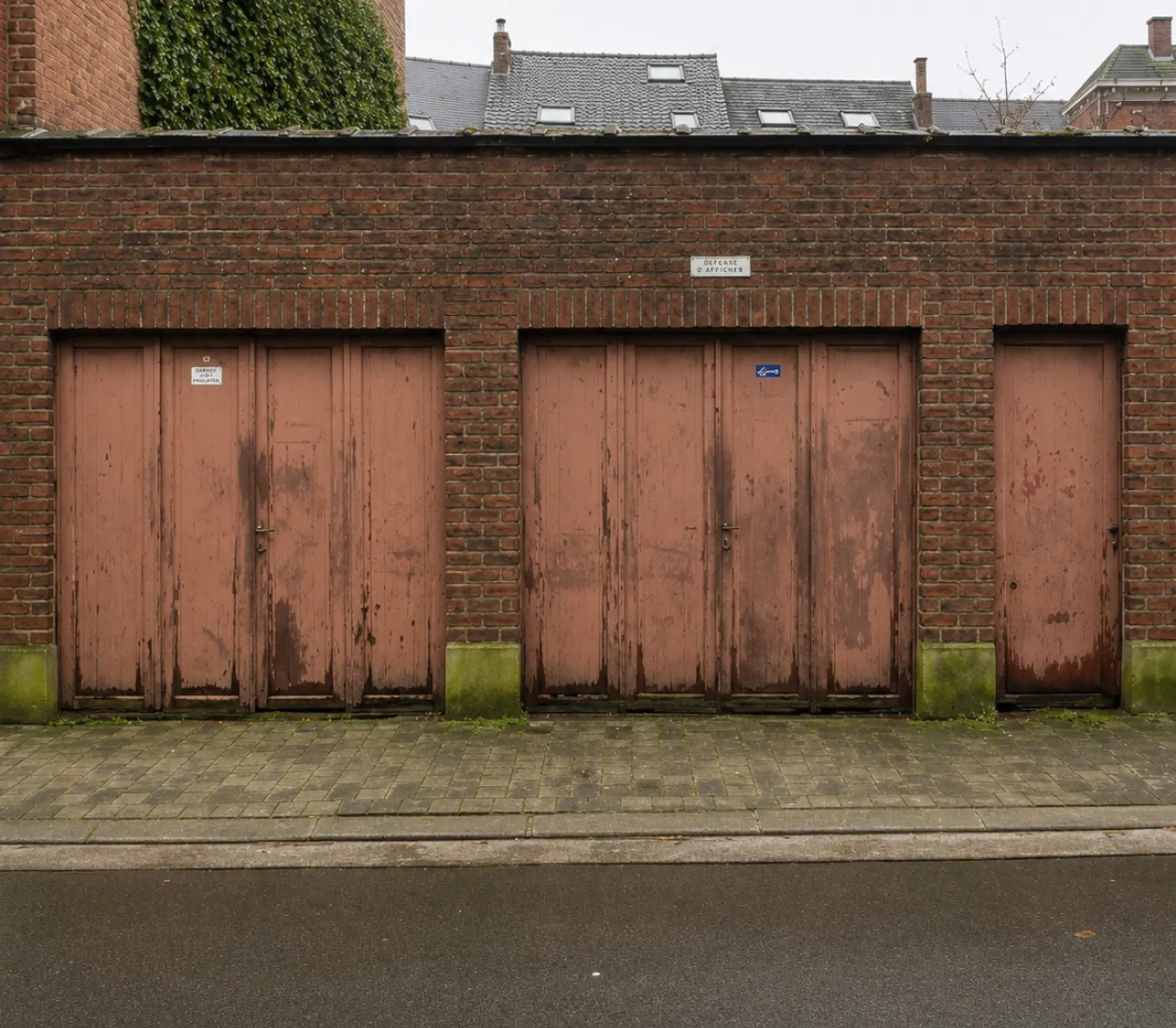 Trois portes en bois usées de couleur rose sur un mur de briques sous un ciel gris.