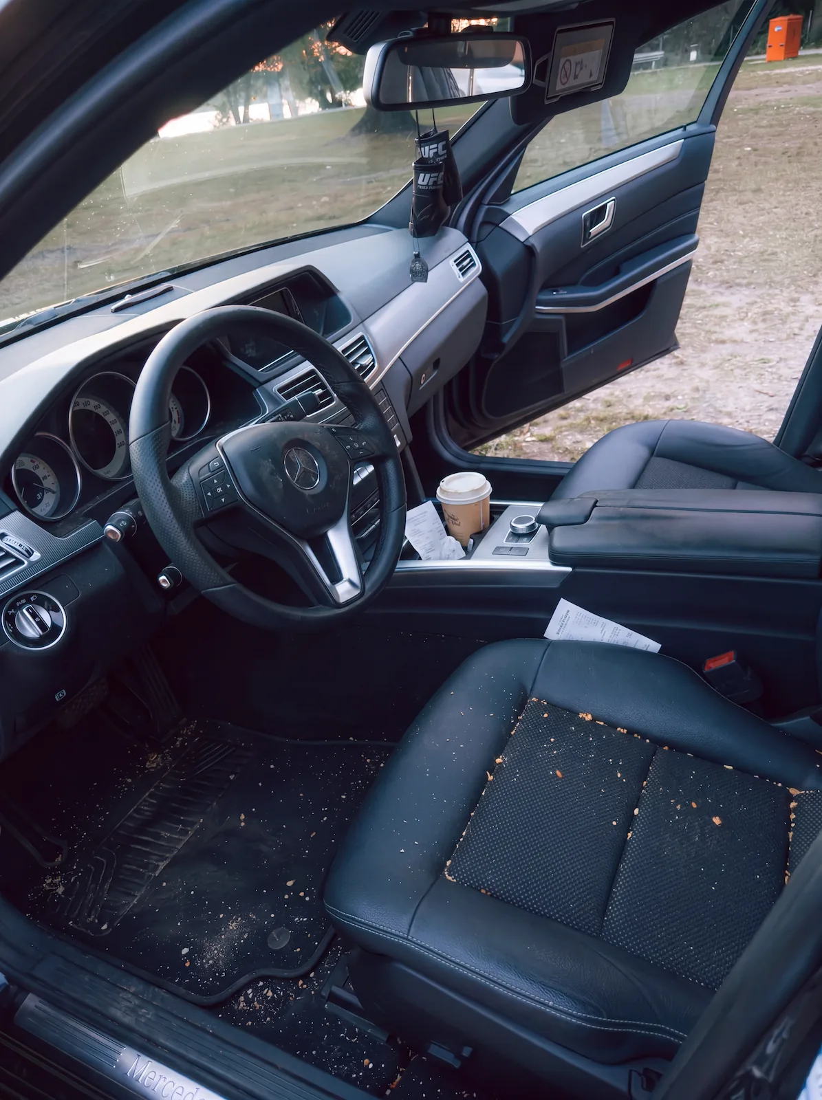 Dirty interior of a Mercedes car with food crumbs on the driver's seat and floor mat, coffee cup, and receipts.