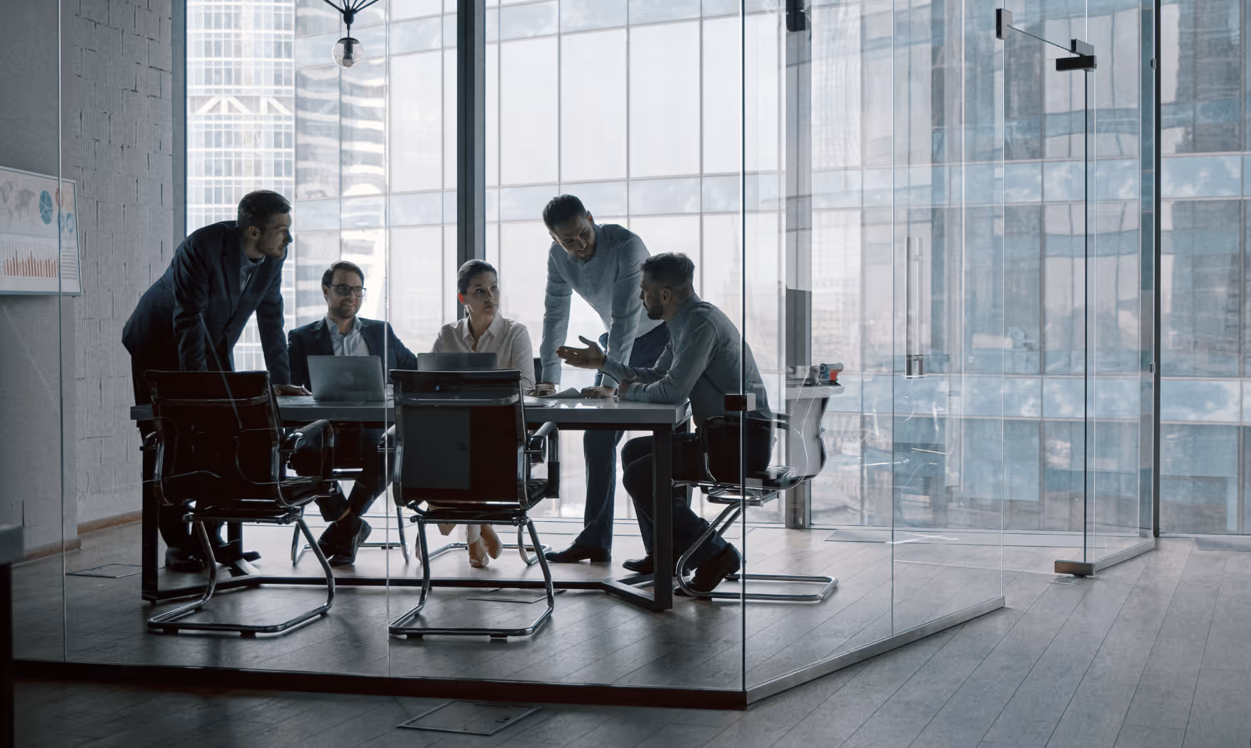 Five business professionals having a discussion around a table in a glass-walled conference room.