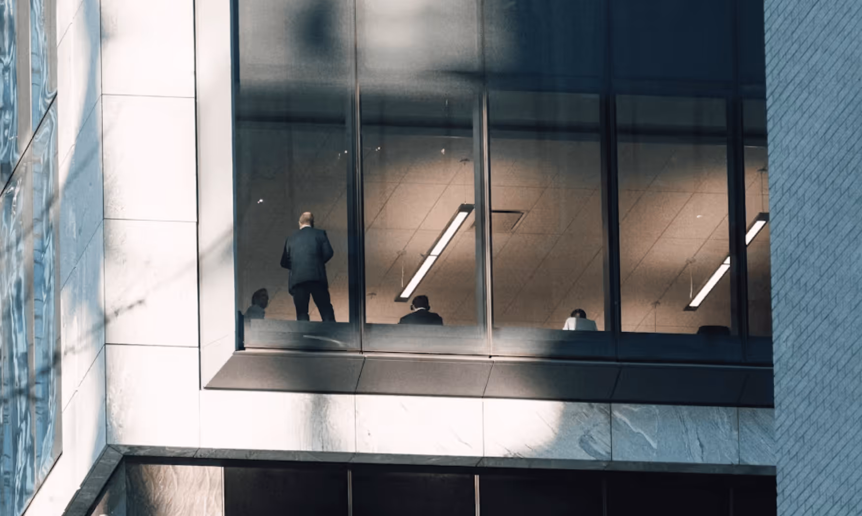 People in business attire seen through office windows in a modern building, some seated and one standing.
