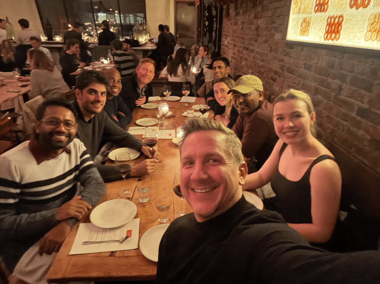 Team members gathered around a long wooden table at a restaurant, smiling for a group selfie with warm lighting and exposed brick walls.
