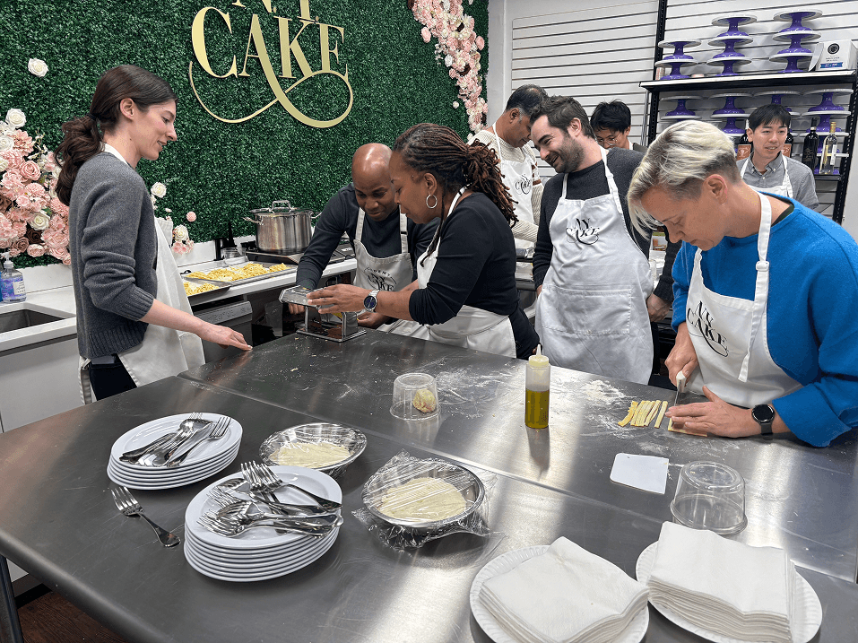 The Foresight team in aprons at a hands-on cooking class, working together at a prep station beneath a floral 'CAKE' sign.