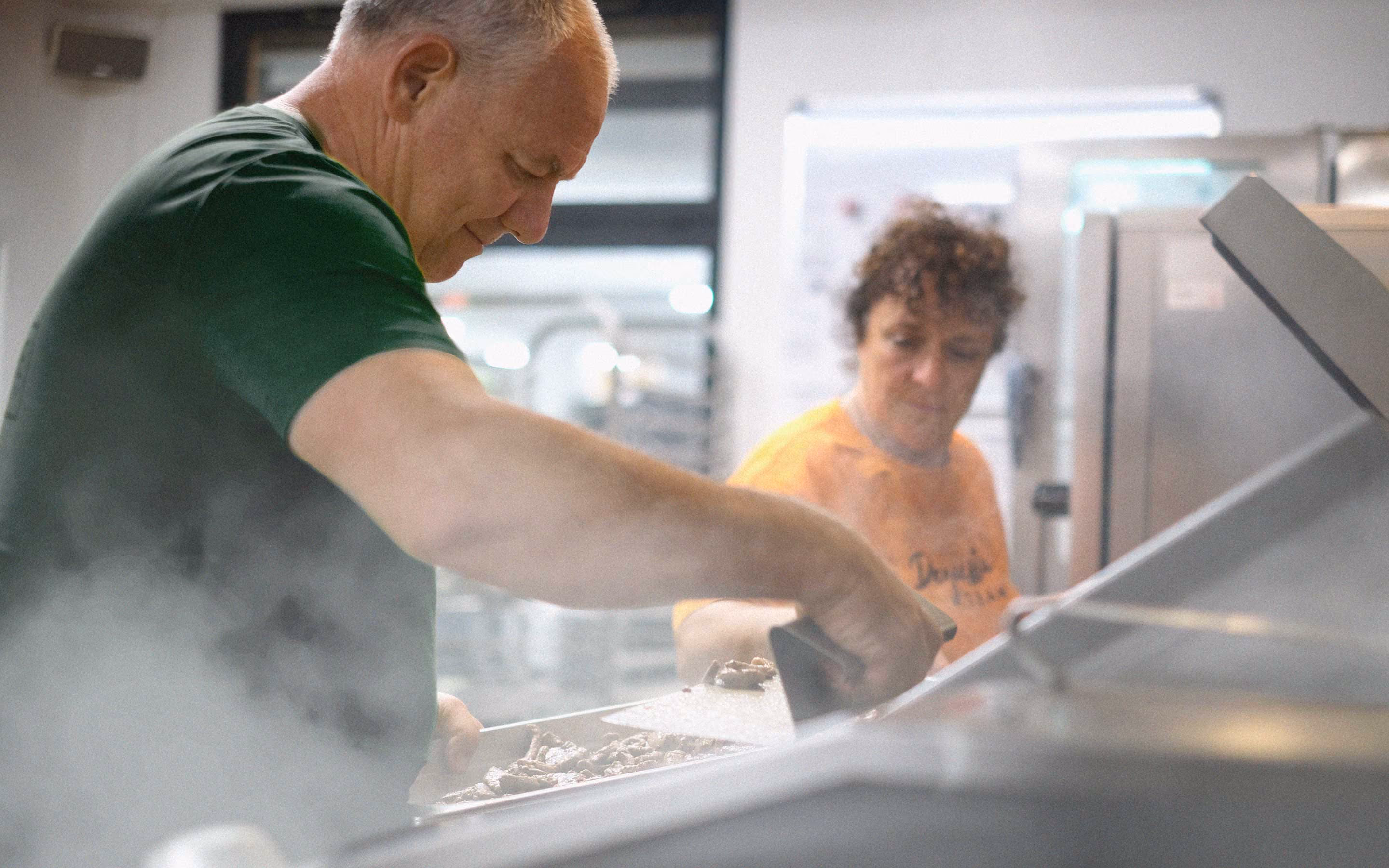 Zwei Personen bereiten dampfendes Essen zusammen in einer professionellen Küche zu.