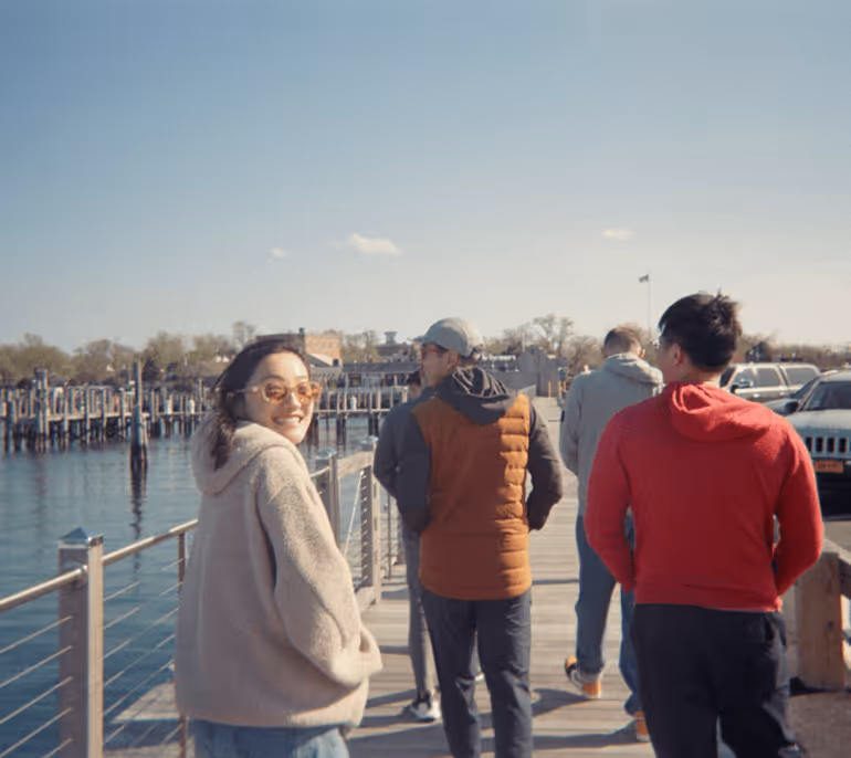 Group of four people walking on a sunny waterfront pier, with one woman smiling back at the camera wearing sunglasses and a beige jacket.