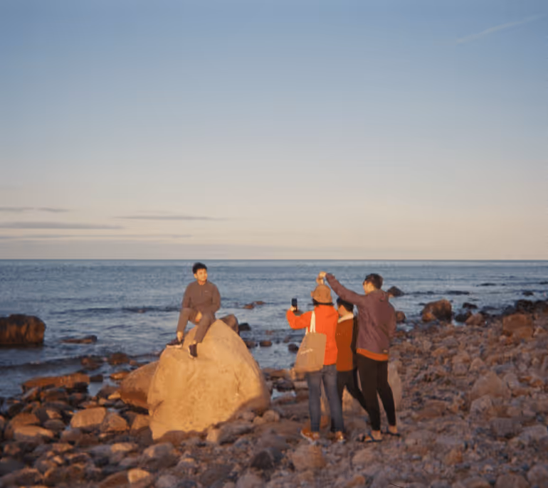 Four people on a rocky beach at sunset with one person sitting on a large rock while another takes a photo with a smartphone.