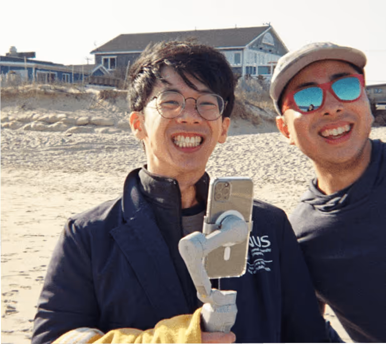 Two smiling men on a sandy beach, one holding a smartphone on a gimbal stabilizer.
