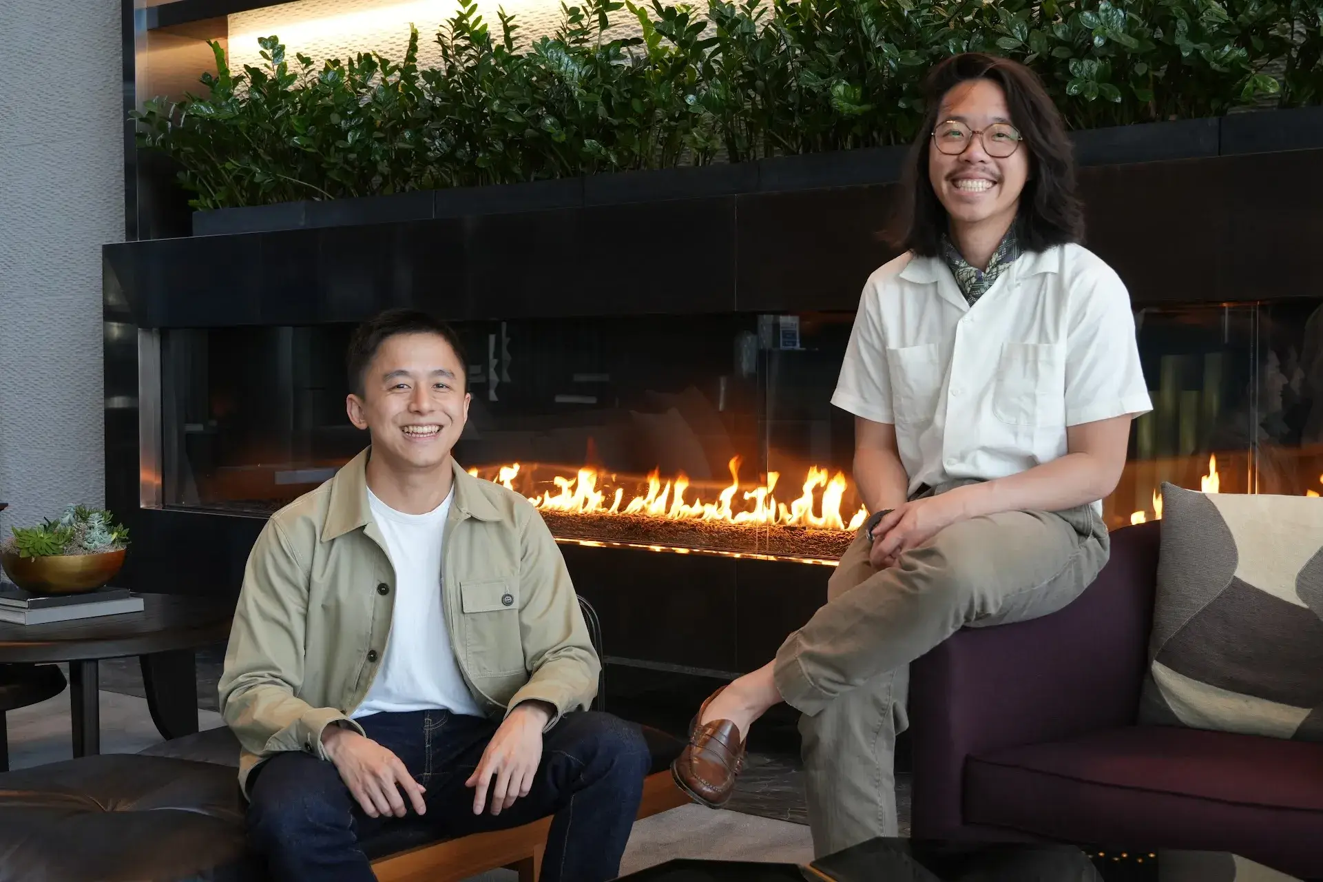 Two men smiling and sitting in a modern lounge area in front of a long fireplace with green plants above it.