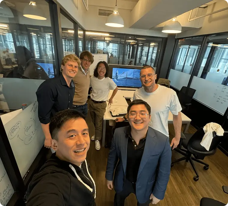 Group of six diverse coworkers smiling and posing for a selfie in a modern office with glass partitions and computer screens.