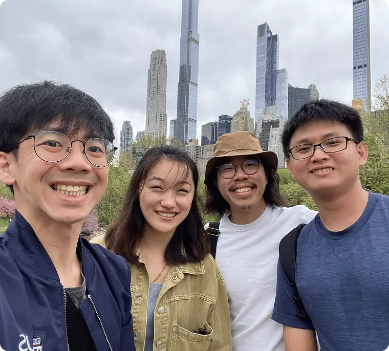 Group of four friends smiling and posing for a selfie in a city park with tall skyscrapers in the background under a cloudy sky.
