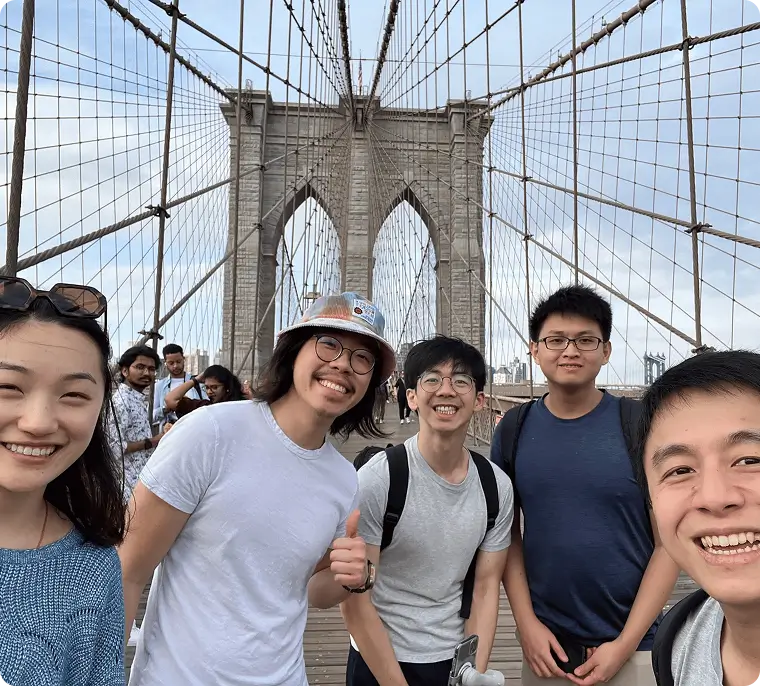 Group of five smiling young adults posing for a selfie on the Brooklyn Bridge with the bridge cables and towers visible.