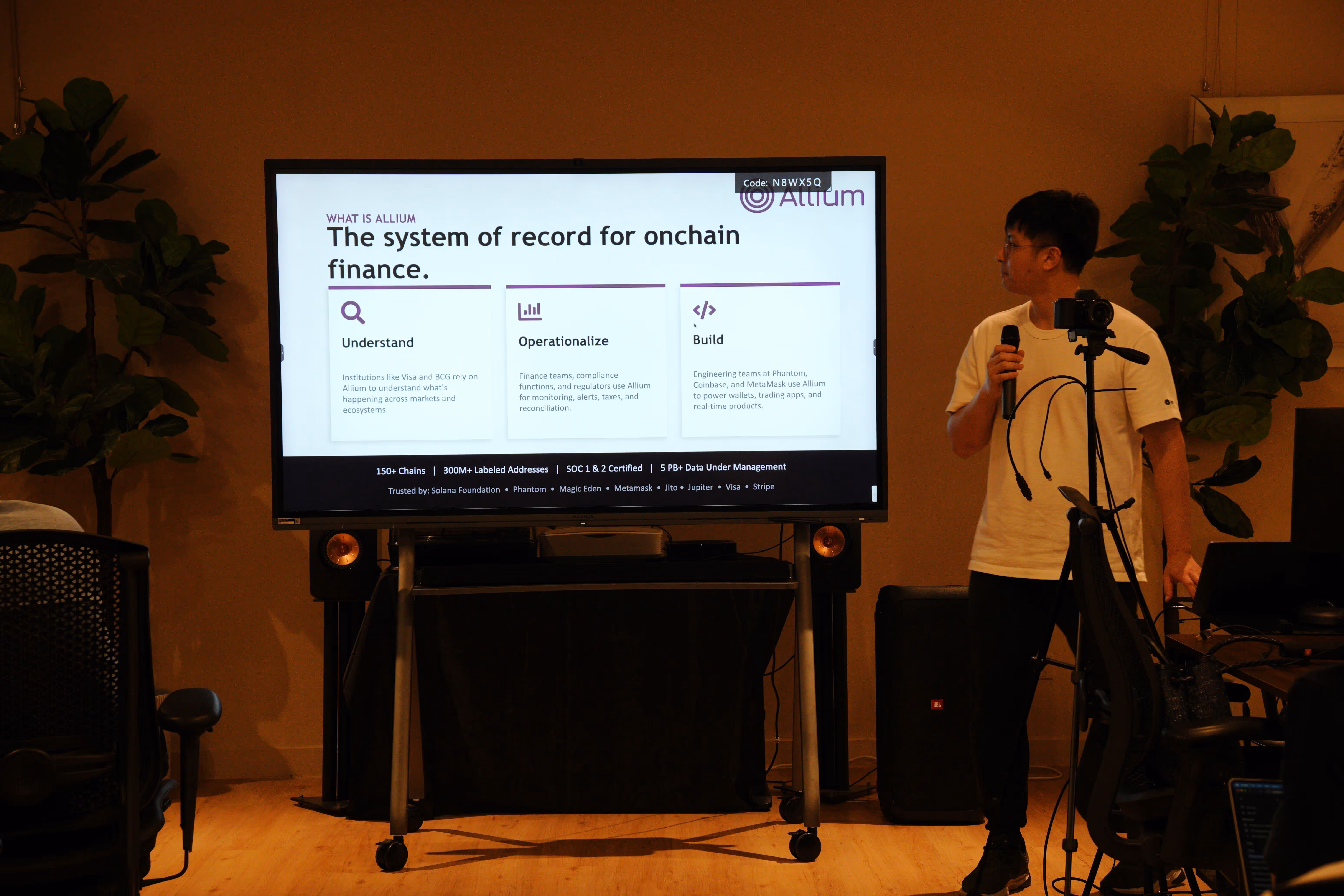Presenter holding a microphone next to a large screen showing a slide about Allium, a system for onchain finance covering Understand, Operationalize, and Build features.