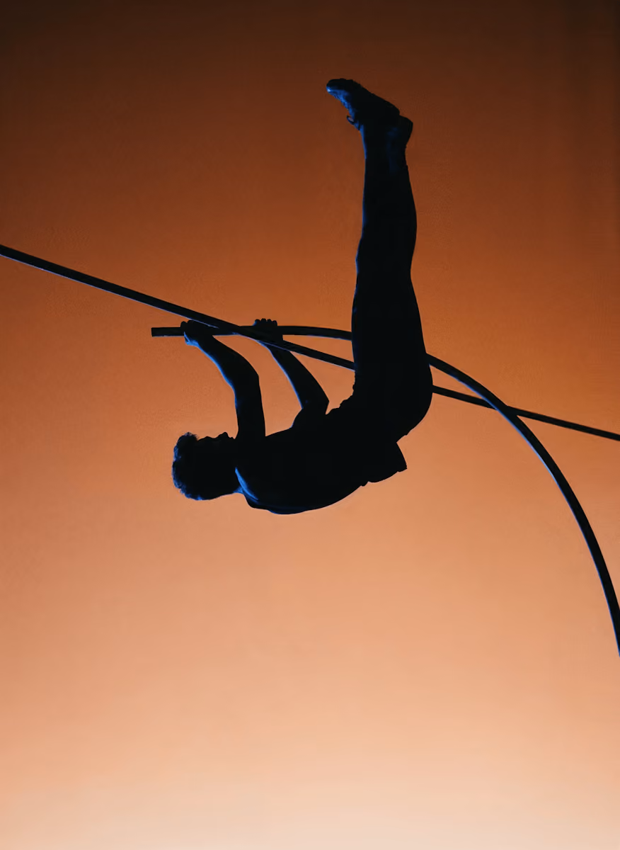 Silhouette of a pole vaulter in mid-air against a gradient orange sky.