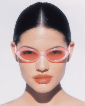 Close-up of a woman with slicked-back hair wearing pink translucent sunglasses and glossy coral lipstick.