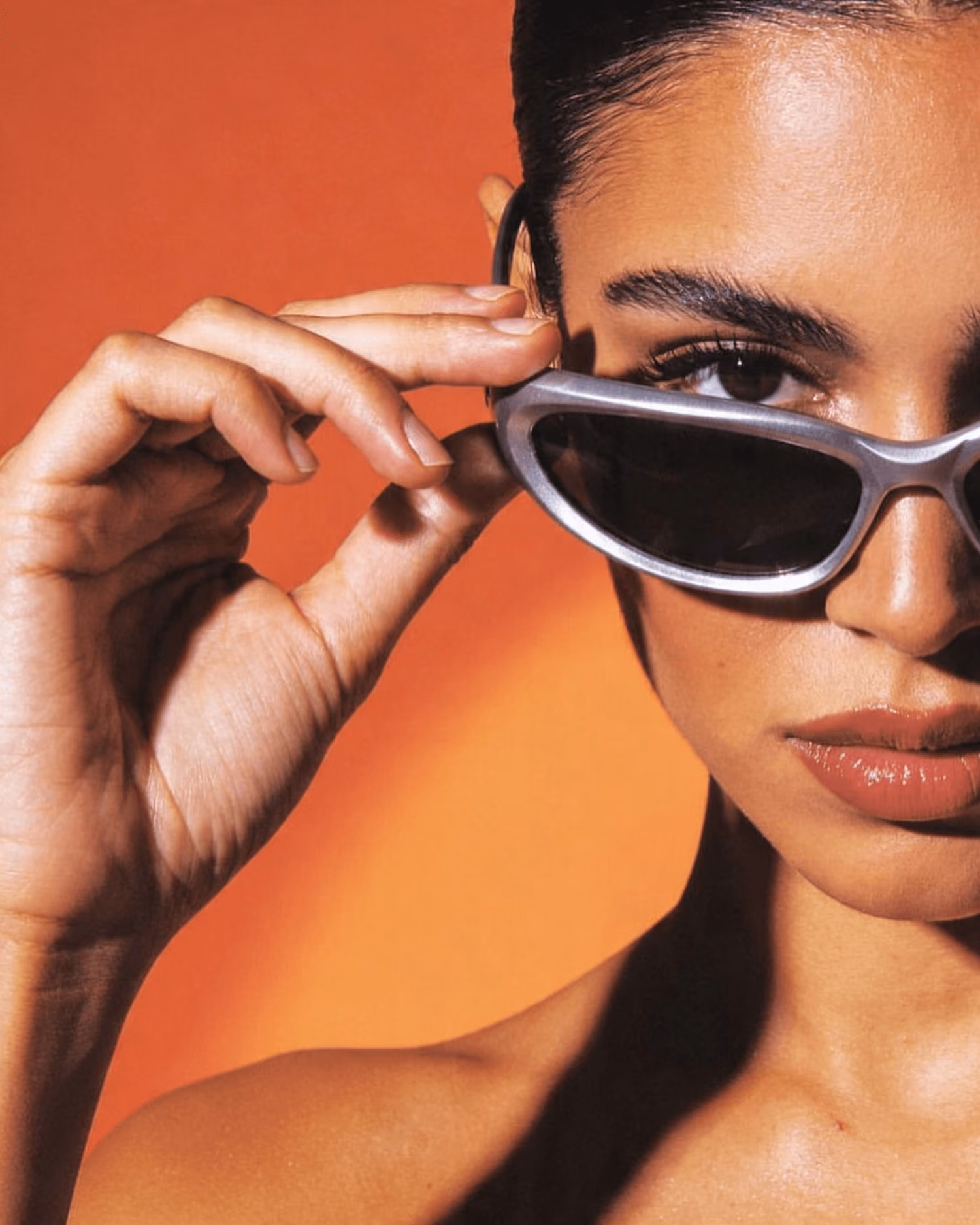 Woman holding silver sunglasses near eye with orange background and confident expression.