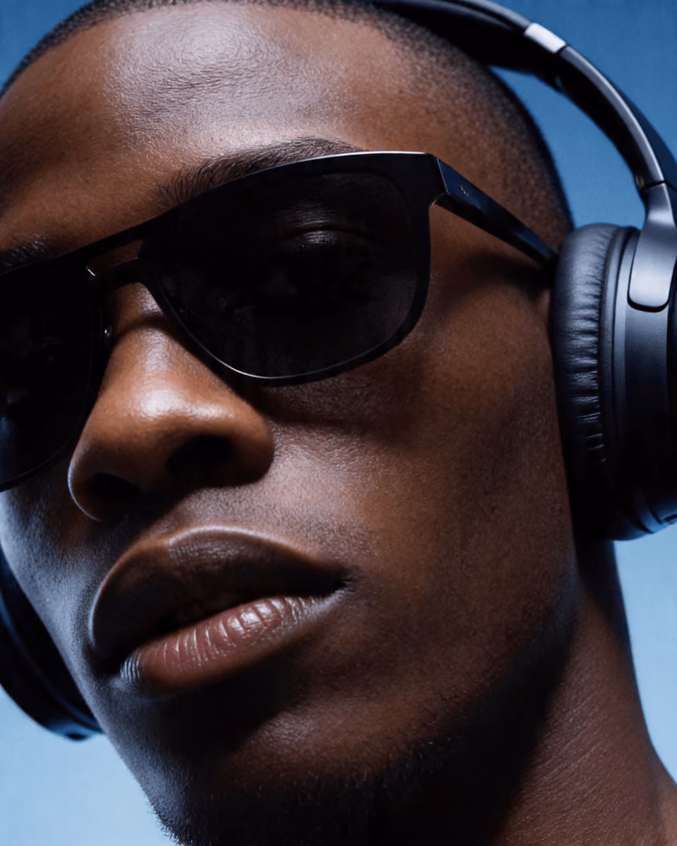 Close-up of a person wearing black sunglasses and over-ear headphones against a blue background.