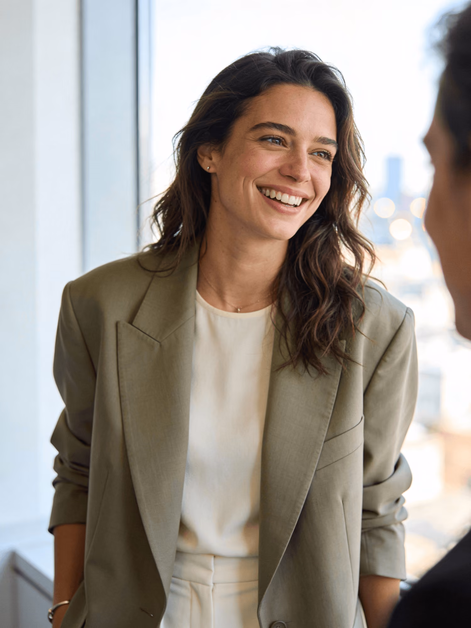 Smiling woman with wavy hair wearing an olive blazer and white top, talking to a person.
