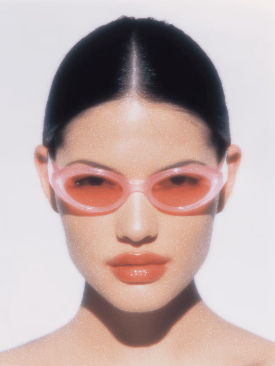 Woman with sleek black hair wearing pink translucent sunglasses and glossy red lips against a white background.