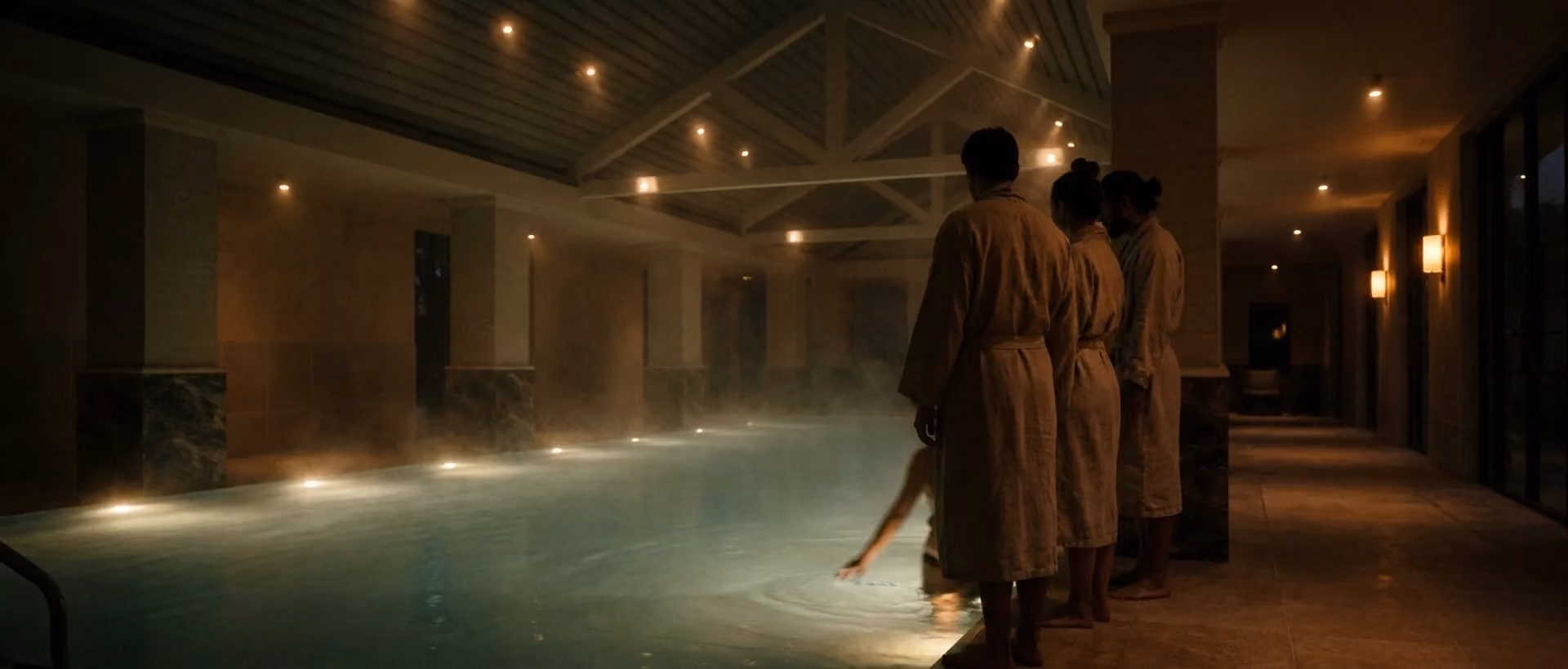 A small group in robes gathered at the edge of the VELA mineral pool at dusk, steam rising from the water