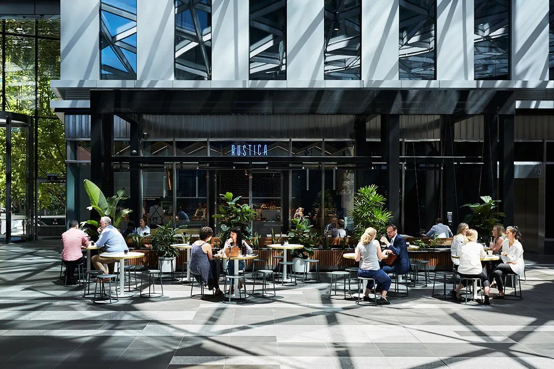 People sitting and talking at tables outside a modern café named Rustica with plants around.