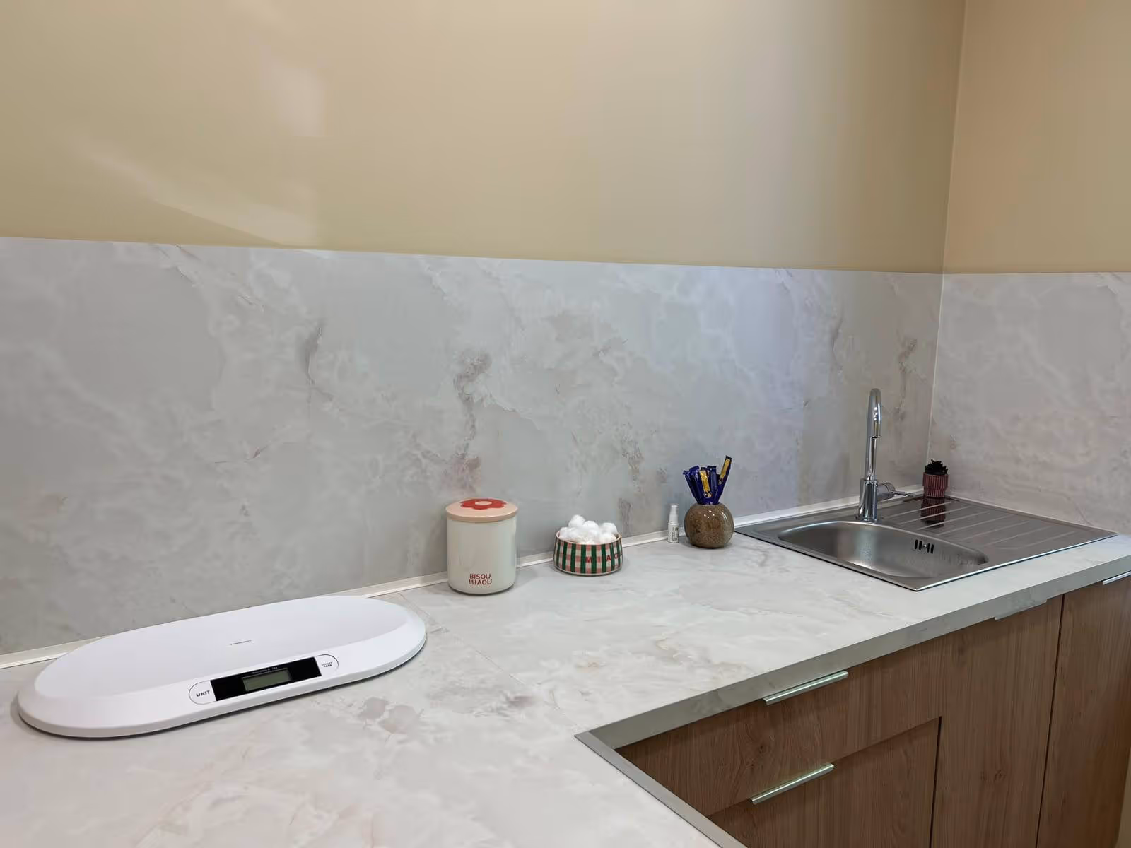 Comptoir de cuisine en marbre avec balance blanche, pot à coton, lavabo en inox et petites décorations.