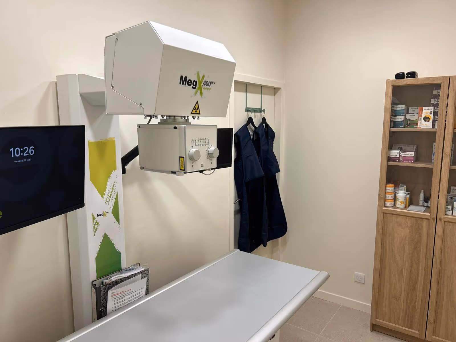 Appareil de radiographie à côté d'une table d'examen, un écran, des tabliers et une armoire à médicaments.