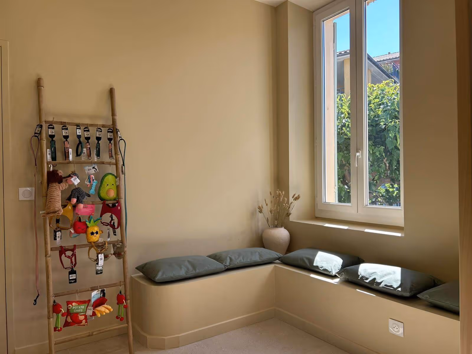 Coin salon beige avec coussins verts, fenêtre ouverte sur jardin et échelle décorative avec jouets.