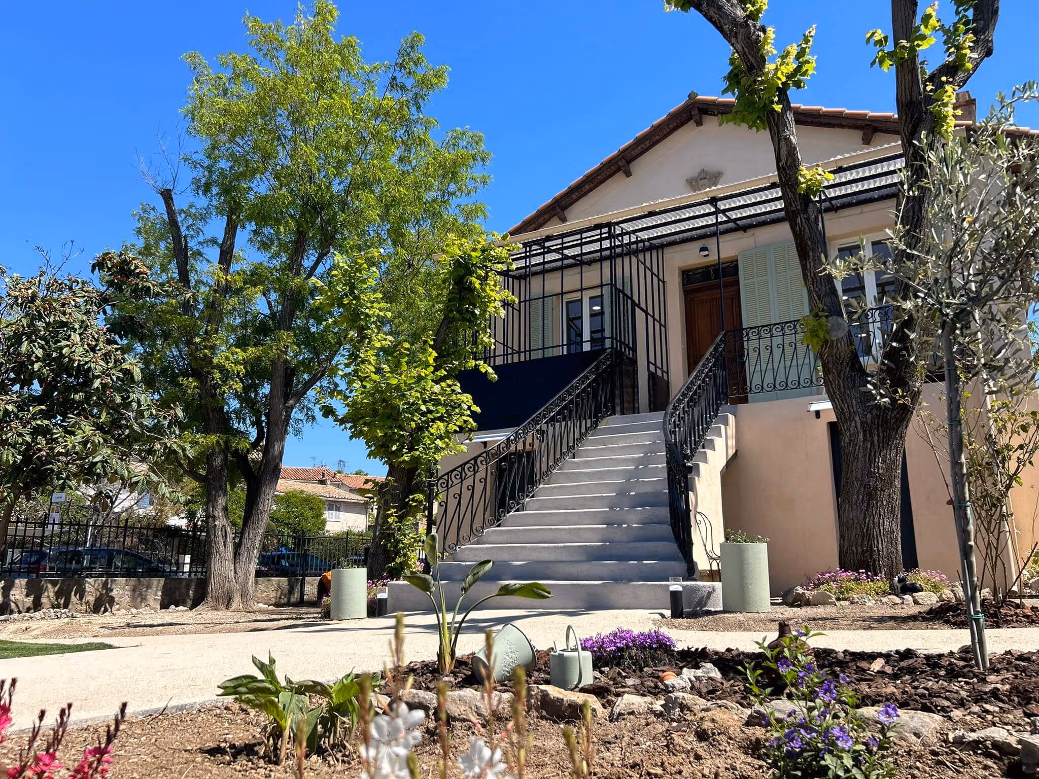 Maison beige avec escalier, jardin fleuri et arbres sous ciel bleu clair.