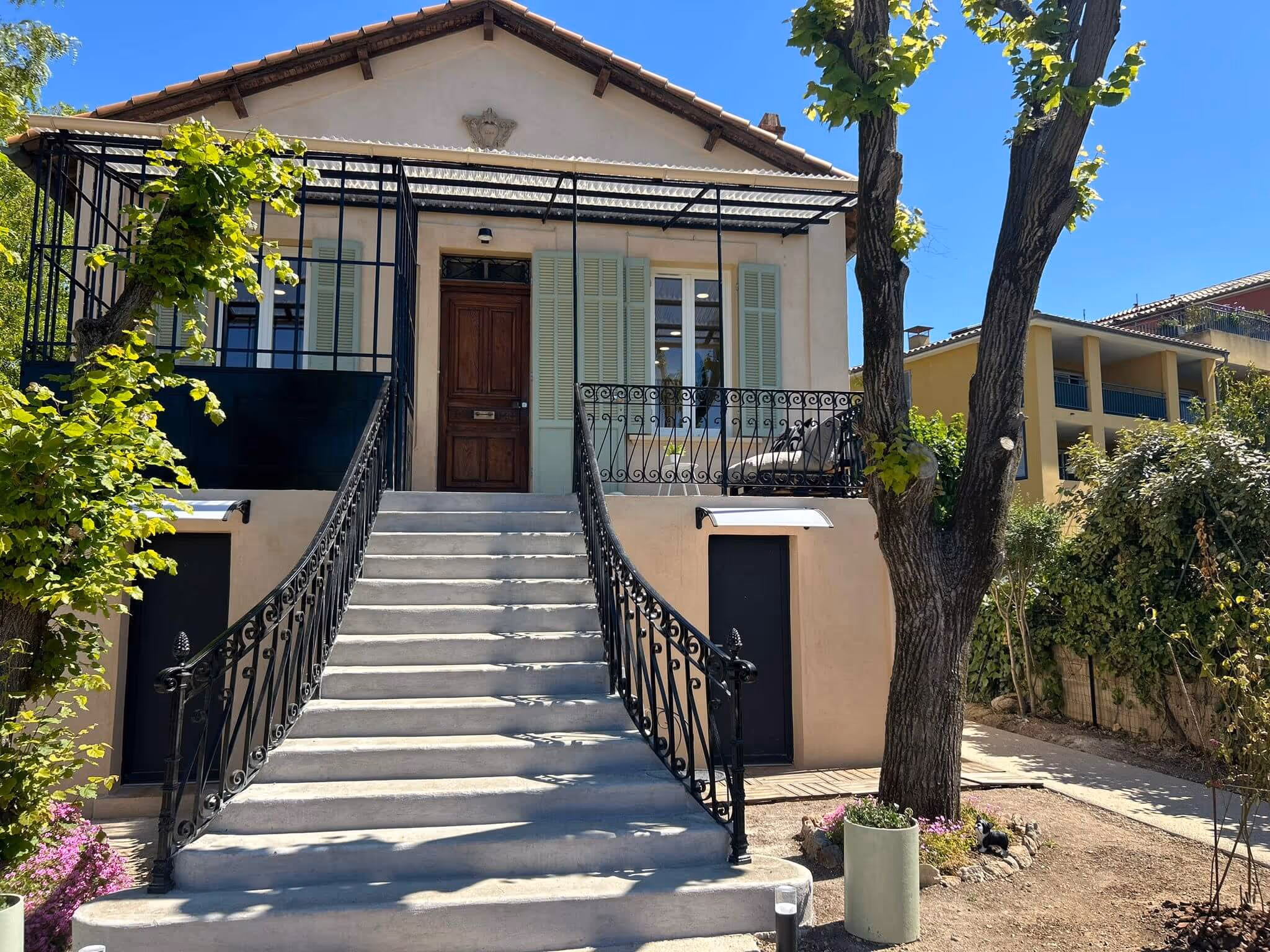 Maison beige avec escalier en béton, porte en bois, volets verts, terrasse et arbres autour.