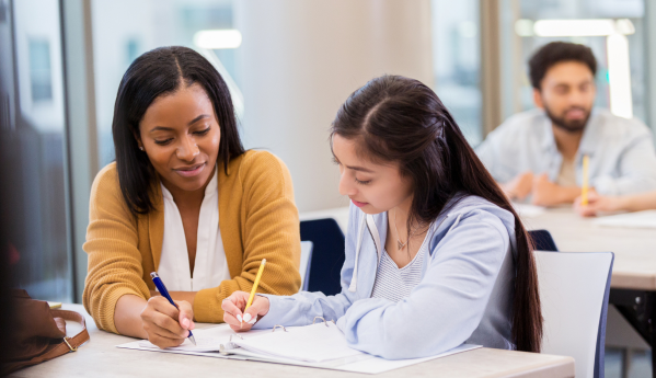 A woman in a brown cardigan helps a student with notes while another student studies in the background.