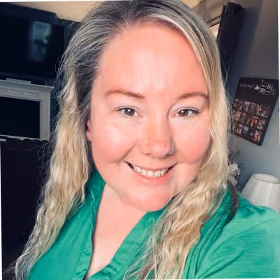 Smiling woman with long blonde hair wearing a green top in a cozy indoor setting.
