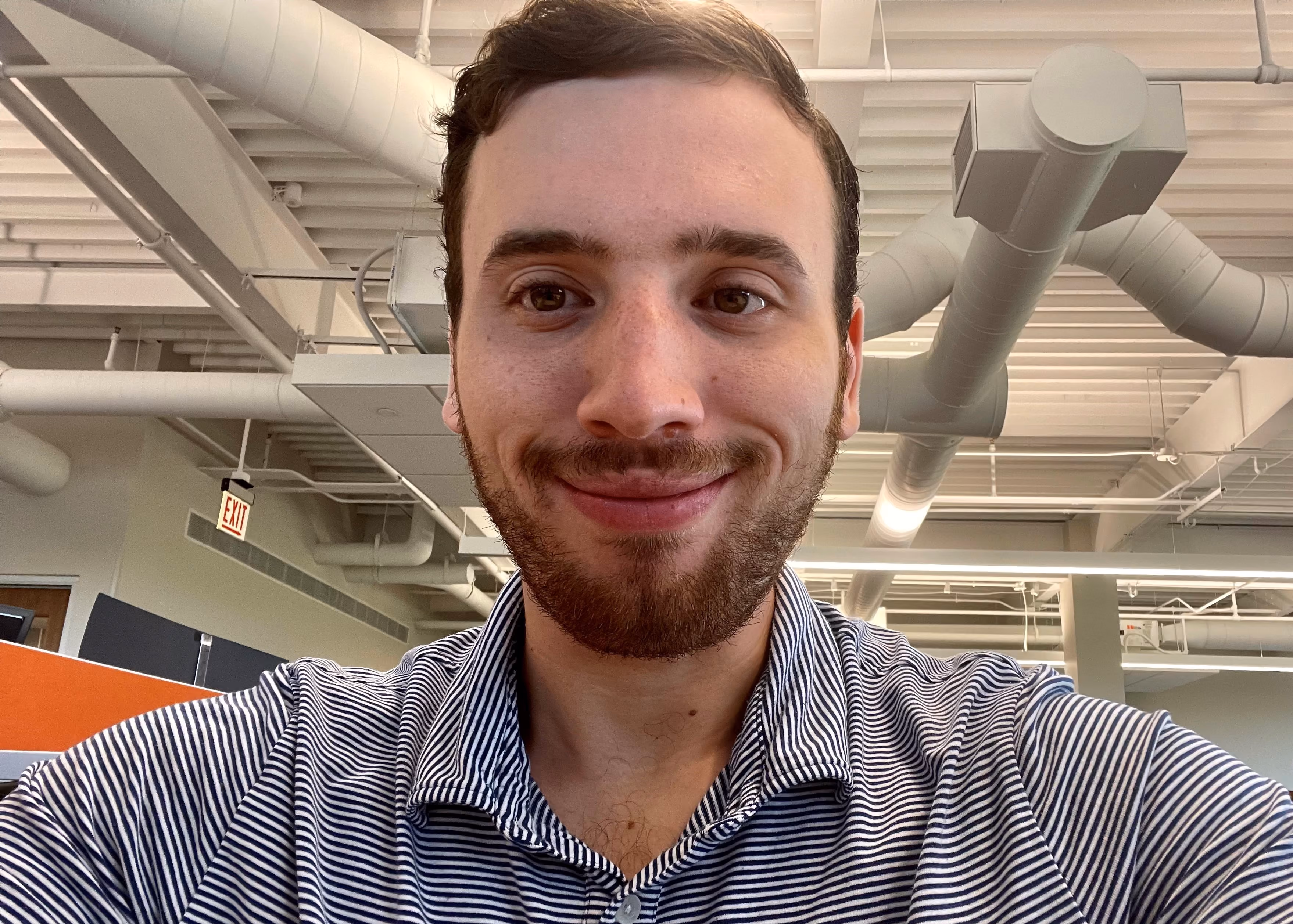 Young man with short brown hair and beard wearing a striped polo shirt in an office with exposed ducts.