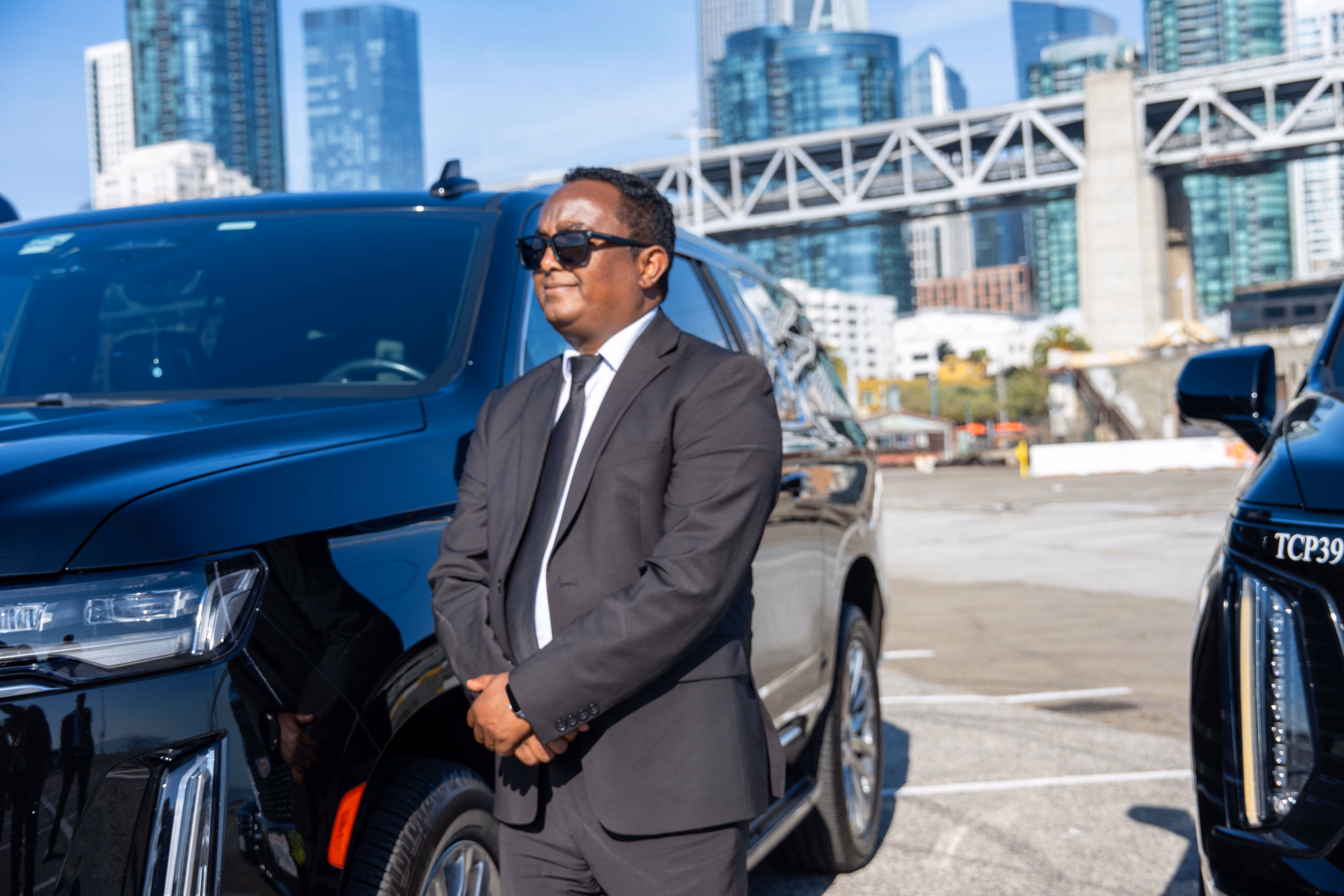 Chauffeur in front of a black Cadillac Escalade with a second SUV and city towers behind